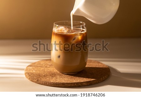 Milk cream is poured into a iced cold brew coffee. Coffee cold cocktail drink with ice and milk in morning sun light. Royalty-Free Stock Photo #1918746206
