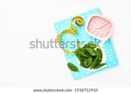 Composition with  spinach, measuring tape, blank paper notebook and pink alarm clock on white background. Mock up. Diet concept, copy space
