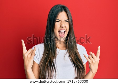 Young brunette woman wearing casual white tshirt over red background shouting with crazy expression doing rock symbol with hands up. music star. heavy concept. 