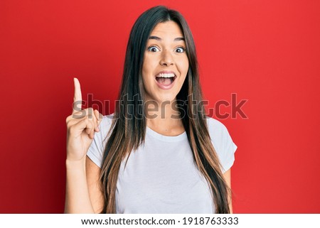 Young brunette woman wearing casual white tshirt over red background pointing finger up with successful idea. exited and happy. number one. 