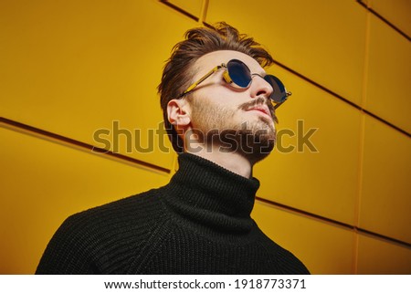 Portrait of a stylish young man in black pullover and round mirror sunglasses looking up on a street by the yellow industrial wall. Male fashion. Urban style. 