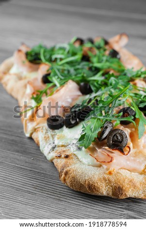 Pinsa romana gourmet italian cuisine on grey wooden background. Scrocchiarella traditional dish. Food delivery from pizzeria. Pinsa with meat, arugula, olives, cheese.