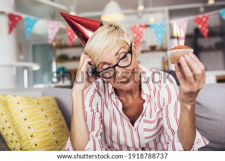 Portrait of senior woman wearing party hat, sitting alone at home on birthday with cake.