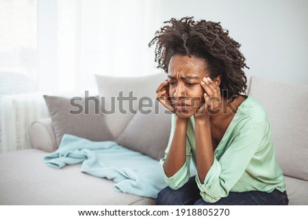 Frustrated woman suffering from the headache while sitting on the sofa at home. Feeling bad. Young woman with headache in home interior. African woman sitting on couch feels unhappy having problems