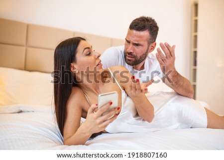 Photo of young couplein bed, woman use smartphone try explain envy angry husband she dont have another man point hand screen feel confused. Jealous husband spying his wife mobile phone