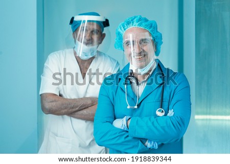 Couple of doctors in scrubs and with all medical protections have their arms crossed and smile with the mask down in a clinical and hospital context.