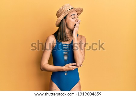 Young brunette girl wearing swimsuit and summer hat bored yawning tired covering mouth with hand. restless and sleepiness. 