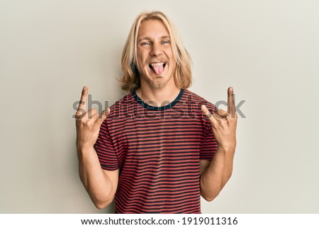 Caucasian young man with long hair doing rock gesture sticking tongue out happy with funny expression. 