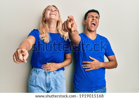 Young interracial couple wearing casual clothes laughing at you, pointing finger to the camera with hand over body, shame expression 