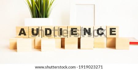 Wooden cubes with letters on a white table. The word is AUDIENCE. White background with photo frame, house plant.