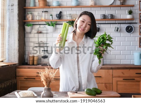 Sporty slim young Asian woman prepares healthy food from green vegetables, fruits and herbs in a bright trendy loft kitchen. The concept of a healthy diet and lifestyle.