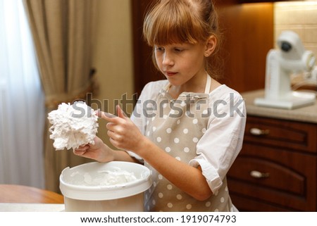A cute redhead girl 10 years old in an apron prepares meringues in the home kitchen. The girl licks her finger, savoring the whipped egg whites.