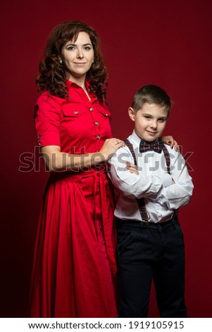 a mother in a dress dress gently hugs her ten-year-old son in a white smart shirt by the shoulders. studio portrait of mom and son on red background with place for text