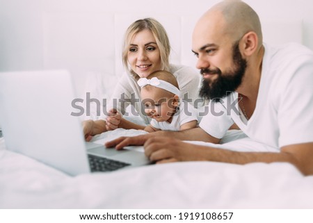A beautiful Caucasian family mom, daughter and bearded dad lie on the bed in the bedroom in the morning and watch cartoons on a laptop. Parents and children under 1 year old.