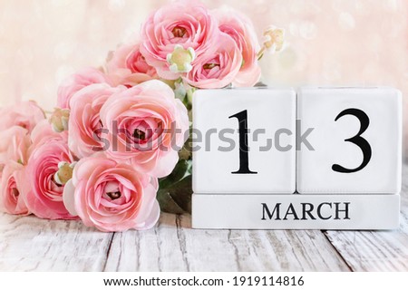White wood calendar blocks with the date March13th and pink ranunculus flowers over a wooden table. Selective focus with blurred background. 
