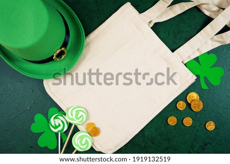 Happy St Patrick's Day natural canvas tote bag, styled with leprechaun hat, shamrocks, and chocolate gold coins, on a textured green background. Mockup. Top view flat lay. Copy space.