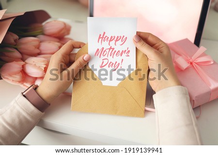 Woman's hands holding a letter in craft envelope. Pink background, Mother's day concept. Tulips flower and pink gift box in background. Womens home desk.