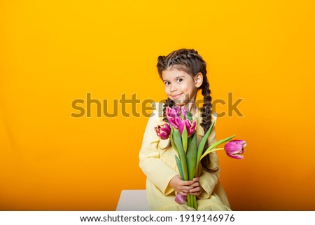 Cute little girl in a yellow dress with tulips on a yellow background. Close-up. Space for text. The concept of spring. The concept of World Women's Day