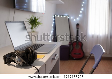 Selective focus of headphones on white desktop and gray laptop, yellow copybook, plant, guitar and mirror defocused. Freelancer workplace concept.