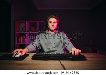 Handsome young man in casual clothes and headset sitting at a table in a room with purple lights and playing video games on the computer with a smile on his face looking at the screen. Royalty-Free Stock Photo #1919169560
