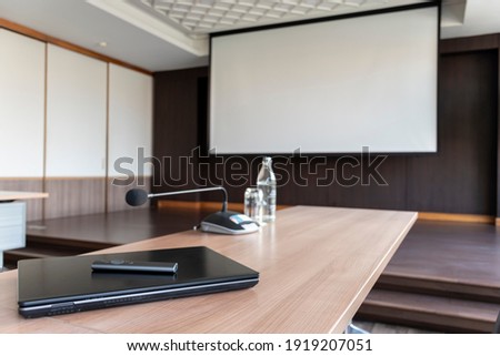 Laptop and remote control slide presentation on table in meeting room with whitesrceen on stage background