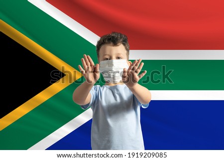 Little white boy in a protective mask on the background of the flag of South African Republic. Makes a stop sign with his hands, stay at home South African Republic.