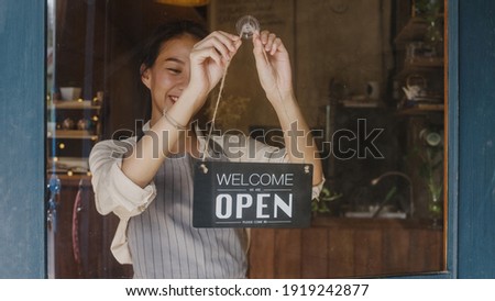 Young Asia manager girl changing a sign from closed to open sign on door cafe looking outside waiting for clients after lockdown. Owner small business, food and drink, business reopen again concept.