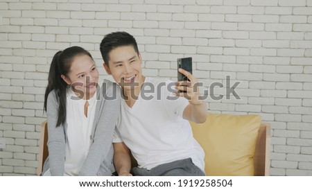 Happy Asian beautiful family couple husband and wife laughing sitting on sofa in the living room taking selfie picture with a smartphone. boyfriend and girlfriend relaxing at home