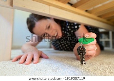 Portrait of a happy beautiful adult woman (female age 30-40) finding home keys under the bed in a domestic house bedroom. Real people. Copy space Royalty-Free Stock Photo #1919307119