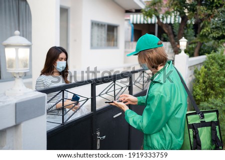 Caucasian delivery man in green uniform giving mobile to Asian woman customer for signing after sending food boxes in plastic bag to her. Both of them wearing a face mask for protective Covid-19