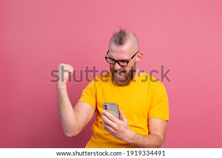Image of screaming young bearded handsome man standing isolated over pink wall background using mobile phone make winner gesture