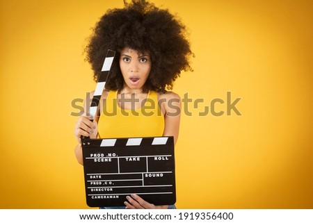Mixed afro american young woman, excited, casual wear, keeping mouth open hold classic black movie clapperboard isolated on orange background. People sincere emotions lifestyle concept. 