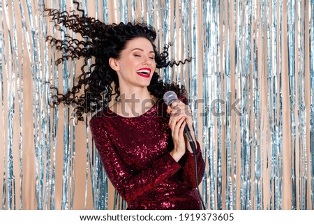Portrait of cheerful pretty girl closed eyes hair flying hand hold mic singing isolated on shiny decorated curtain