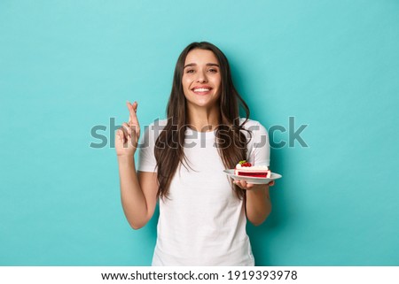 Image of happy and excited brunette woman, celebrating her birthday, cross fingers while making wish, holding b-day cake with lit candle