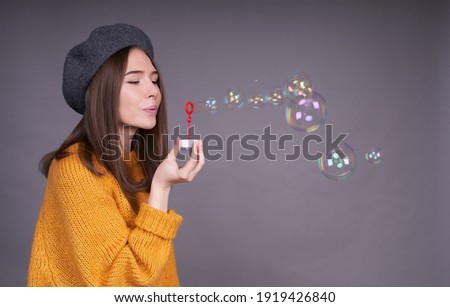 A beautiful cute young Caucasian girl in a bright yellow sweater, a gray beret, with a stylish bob hairstyle blowing soap bubbles, stands sideways on a gray background.