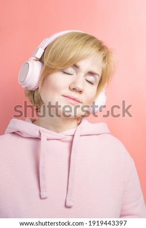 Caucasian girl in a pink sweatshirt and with pink headphones on a background similar in tone. Concept for music, podcasts, audiobooks. World Music Day, World Rock-n-roll Day.