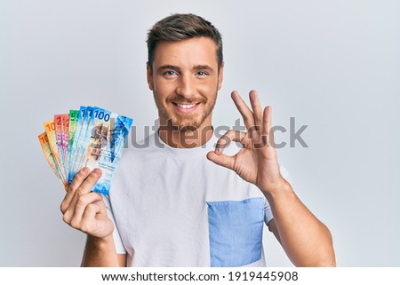 Handsome caucasian man holding swiss franc banknotes doing ok sign with fingers, smiling friendly gesturing excellent symbol 