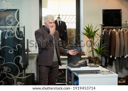 A mature man with gray hair and a sporty physique is holding a credit card to pay for purchases in a clothing store. A customer with a beard is massaging his face in a boutique