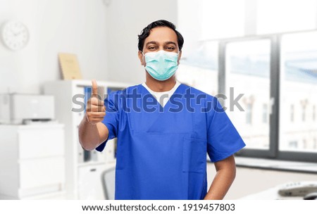 healthcare, profession and medicine concept - indian doctor or male nurse in blue uniform and face mask for protection from virus showing thumbs up over medical office at hospital on background