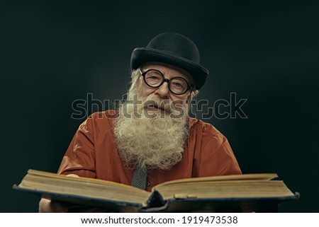 An intelligent old man in glasses and bowler with a long gray beard holds an old big book and looks at the camera. Old age wisdom. Royalty-Free Stock Photo #1919473538