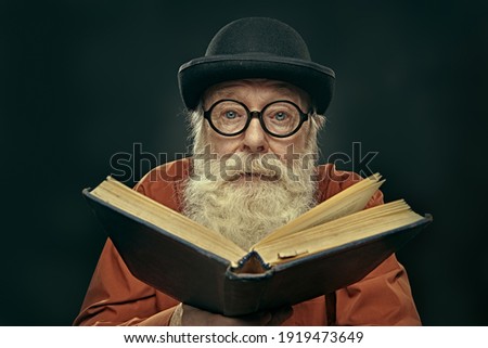 Portrait of an intelligent old man in glasses and bowler with a long gray beard holding an old big book and looking at the camera. Old age wisdom. Royalty-Free Stock Photo #1919473649