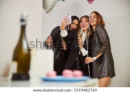 Front view of three joyous ladies with balloons and champagne flutes being photographed against the wall Royalty-Free Stock Photo #1919482769