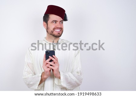 young handsome Caucasian man wearing Arab djellaba and Fez hat over white wall hold telephone hands read good youth news look empty space advert