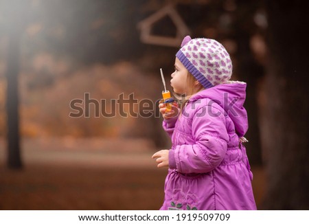 Beautiful little blonde girl, has pretty face, happy emotions, dressed in lilac coat. Plays soap bubble blower. Child portrait. Lifestyle instagram concept. Autumn time. Fashion kid style.