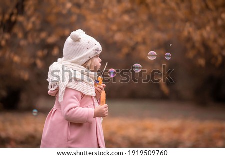 Beautiful little blonde girl, has fun smiling face, happy emotions, dressed in pink coat. Plays soap bubble blower. Child portrait. Lifestyle instagram concept. Autumn time. Fashion kid style.