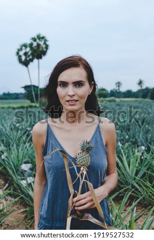 Young Beautiful Woman wearing overalls is standing in a Pineapple Field among Pineapples (Prachuap Khiri Khan, Thailand). Woman with Pineapple. Summer Fashion.