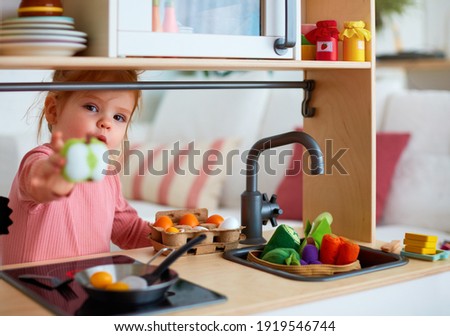 cute toddler baby girl playing on toy kitchen at home, roasting eggs and treat you with apple slice, let's share Royalty-Free Stock Photo #1919546744