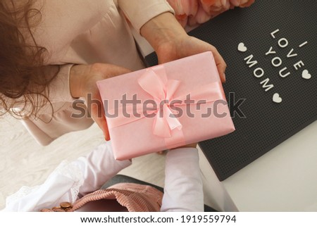 Child's hands hold beautiful pink gift box with ribbon. Pink tulip flower and sign i love you mom in background. Top view, close-up. Preparing for the holidays.