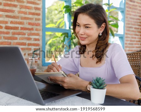Cute beautiful middle-aged Asian woman sitting in shop and using a pencil to write on a paper notebook with a laptop computer. Work from anywhere concept.