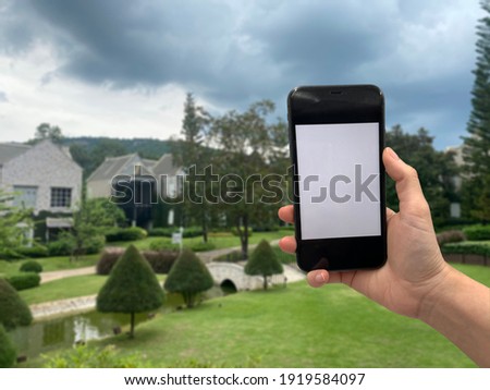 
Human hand is holding phone for taking photo with blur garden , bridge and  European building house  background